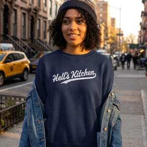 May include: Navy blue sweatshirt with the text "Hell's Kitchen" in white script. The sweatshirt is worn with a denim jacket and a striped beanie. The image is taken outdoors on a city street.