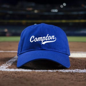 May include: A royal blue baseball cap with a white embroidered cursive script that reads "Compton". The cap is sitting on a baseball field.