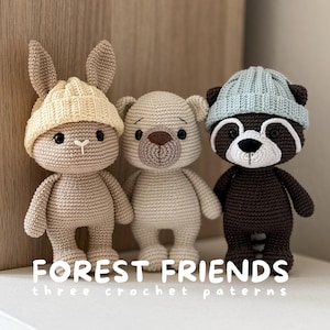 Puede incluir: Tres animales de peluche tejidos a ganchillo que incluyen un conejo beige con gorro amarillo, un oso color crema y un mapache marrón oscuro con gorro azul claro. El texto FOREST FRIENDS three crochet paterns está impreso en blanco.