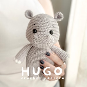 Pode incluir: Um brinquedo de hipopótamo em crochê cinza-claro com olhos de segurança pretos e pequenas orelhas arredondadas. O texto HUGO crochet pattern está impresso em letras brancas na parte inferior da imagem, centralizado sobre o corpo do brinquedo.