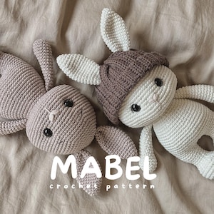 May include: Two handmade crochet dolls shaped like rabbits. One is solid light brown, and the other is cream-colored with a dark brown ribbed beanie hat. Both have black plastic eyes and stitched noses. The text MABEL crochet pattern is visible.