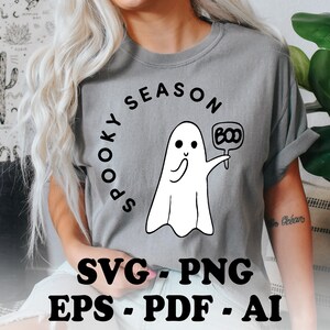Puede incluir: Una camiseta gris con un fantasma blanco sosteniendo un cartel que dice "BOO". El texto "SPOOKY SEASON" está en un círculo alrededor del fantasma. La camiseta está disponible en los formatos SVG, PNG, EPS, PDF y AI.