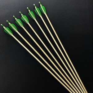 May include: A set of eight wooden arrows with green fletching and black tips. The arrows are arranged in a fan shape on a black background.