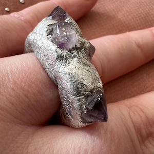 May include: A silver ring featuring three amethyst crystals set into the band. The ring has a textured finish and a distinctive design.