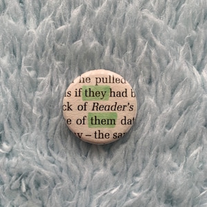 Op de afbeelding: Een witte knop met een citaat uit het boek "De Lezer" van Bernhard Schlink. Het citaat luidt "is if they had b ck of Reader's e of them dat v - the sar".
