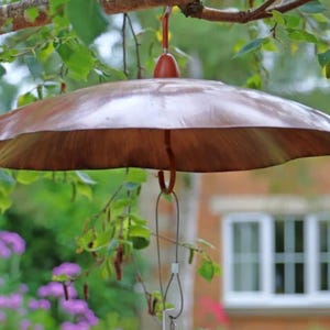 May include: A brown, umbrella-shaped bird feeder suspended from a tree branch. The feeder has a rounded top and a decorative, scalloped edge. The background features green foliage, purple flowers, and a house with white windows.