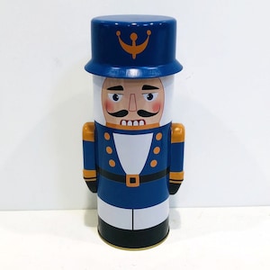 May include: A blue and gold nutcracker tin with a white face and a gold mustache. The tin is shaped like a soldier and has a blue hat with a gold star.