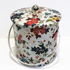 May include: A white metal ice bucket with a floral pattern in shades of red, blue, yellow, and green. The bucket has a lid with a silver knob.