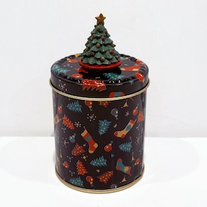 May include: A round, dark brown tin with a Christmas-themed pattern of trees, socks, and ornaments. The lid is topped with a green Christmas tree figurine with a gold star. The tin has a gold rim.