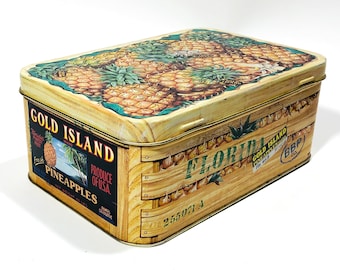 May include: A vintage tin box with a wood-grain design and a picture of pineapples on the lid. The box is labeled "Gold Island" and "Florida" with a sticker that says "Gold Island Pineapple Products".