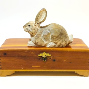 May include: A wooden rabbit figurine sits on top of a cedar wood box with a brass latch. The rabbit is carved in a detailed, layered style and is painted white.