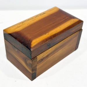 May include: A small wooden box with a hinged lid. The box is made of cedar wood and has a natural wood finish. The lid is engraved with the words "Made in the USA".