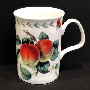 May include: White ceramic mug with a red apple and green leaves design. The mug has a handle and is decorated with a floral pattern.