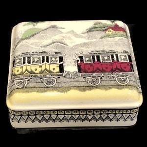 May include: A vintage ceramic trinket box with a colorful illustration of a train traveling through a landscape. The box has a yellow, black, and red train with a white background. The box has a decorative border around the bottom.
