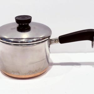 May include: A silver saucepan with a copper bottom and a black handle. The saucepan has a lid with a black knob.