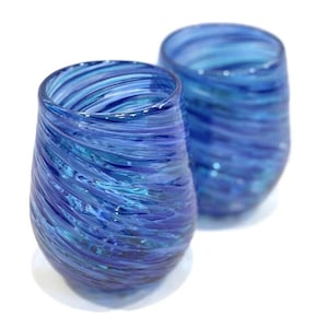 May include: Two stemless, blue glass tumblers with a swirling, textured design. The glasses have a rounded shape and are made of transparent glass, allowing light to pass through. The color transitions from a deep blue to lighter shades.