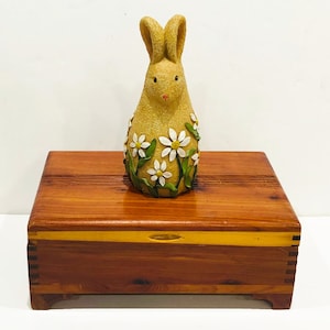 May include: A wooden box with a hinged lid and a yellow bunny figurine with white flowers on it. The box is made of cedar wood and has a natural finish.
