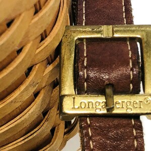 May include: A close-up of a brown leather strap with a gold buckle. The buckle has the words "Longaberger" engraved on it.