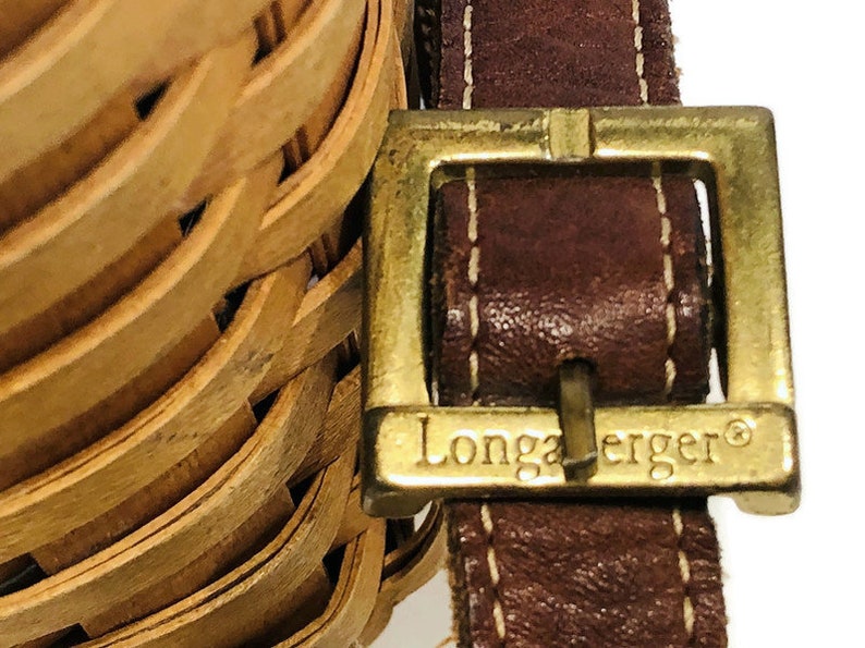 May include: A close-up of a brown leather strap with a gold buckle. The buckle has the words "Longaberger" engraved on it.