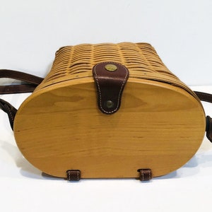 May include: A brown leather-trimmed wicker basket with a wooden bottom and a brown leather strap. The basket is closed and the strap is buckled.