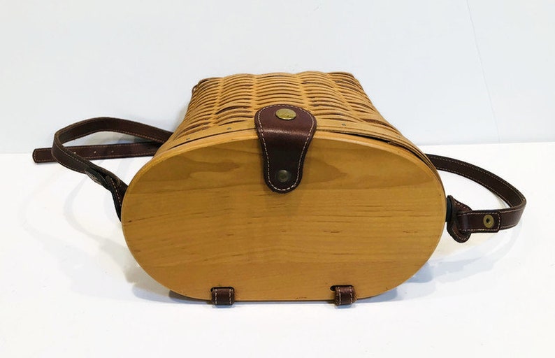 May include: A brown leather-trimmed wicker basket with a wooden bottom and a brown leather strap. The basket is closed and the strap is buckled.