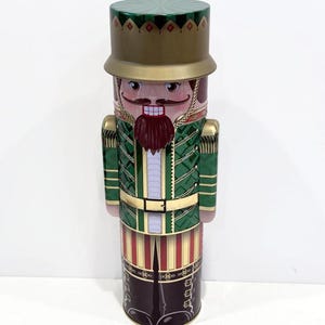 May include: A decorative tin shaped like a nutcracker soldier. The tin features a green and gold uniform with a red beard and a stern expression. The hat is green with gold trim, and the base of the tin is dark brown with red and white stripes.