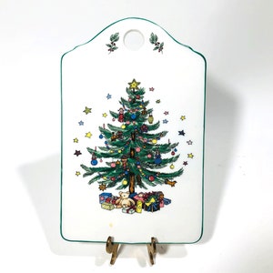 May include: White ceramic Christmas tree cutting board with a green border. The board features a green Christmas tree with colorful ornaments, presents, and a teddy bear at the base. The tree is surrounded by small stars.