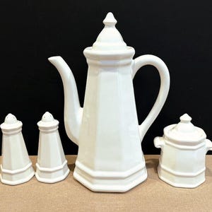 May include: A white ceramic coffee set featuring a tall coffee pot, a sugar bowl with a lid, and two salt and pepper shakers. The set has a classic, elegant design, perfect for a kitchen or dining room.