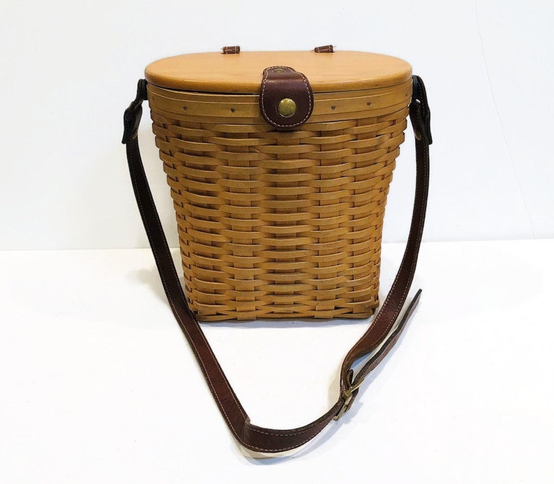 May include: A brown wicker basket with a lid and a brown leather strap. The basket is made of woven wicker and has a natural wood finish.