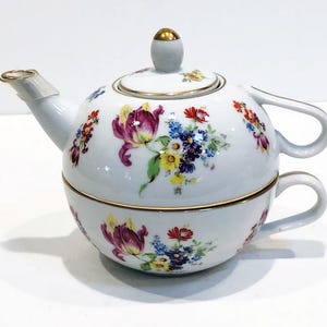 May include: A white porcelain teapot with a floral design. The teapot has a gold trim and a lid with a knob. The teapot is sitting on a white surface.