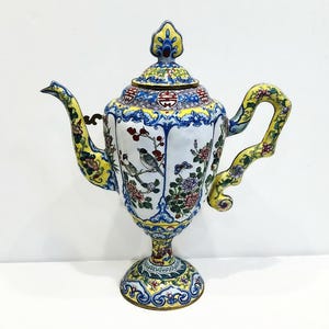 May include: A white porcelain teapot with a yellow, blue, and red floral design. The teapot has a round body with a tall spout and a handle that curves up and over the top. The teapot sits on a round base with a floral design.