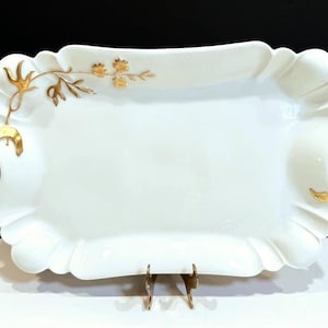 May include: A rectangular white porcelain serving tray with scalloped edges and two gold-toned handles. The tray features a delicate gold floral branch design on the upper left corner and a small gold leaf accent on the lower right side.