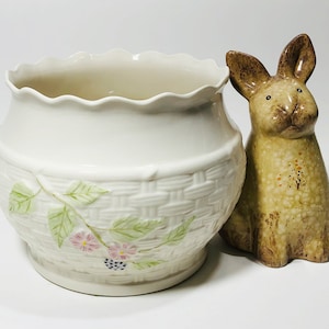 May include: A white ceramic planter with a woven basket design and floral accents. The planter is next to a brown ceramic bunny figurine.