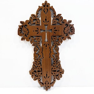 May include: A wooden cross with intricate, openwork floral and scroll designs. The cross is a warm brown color and features a smaller cross at the center. The edges are detailed with carved patterns, creating a decorative religious item.