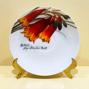 May include: White ceramic saucer with a floral design featuring red and orange flowers. The saucer has a gold metal stand and the text "Botanica Large Christmas Bells" is printed on the bottom.