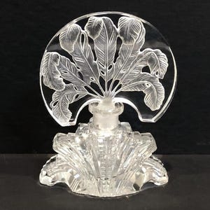 May include: A clear glass perfume bottle with a decorative stopper featuring a fan of etched feathers. The bottle has a faceted design and a rounded base.