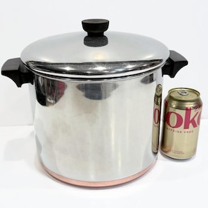 May include: A large stainless steel stockpot with a copper-colored base, two black side handles, and a matching lid with a black knob. A standard 12-ounce soda can is placed next to the pot to provide a visual reference for its overall size.
