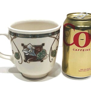 May include: A white ceramic mug with a floral design featuring a stylized flower with green leaves and a brown stem. The mug has a gold trim around the top and a handle on the left side. The mug is sitting on a white surface next to a can of Coca-Cola.