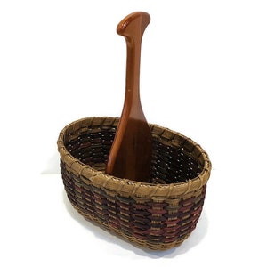 May include: A woven basket with a wooden handle. The basket is oval shaped and has a brown, red, and green pattern.