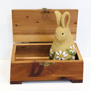 May include: A wooden box with a hinged lid and a brass latch. The box is open and a ceramic bunny figurine with white flowers is inside.