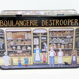 May include: A rectangular black tin with a colorful illustration of a French bakery, "BOULANGERIE DESTROOPER", with various baked goods, people, and text. The tin has a hinged lid and a white background.