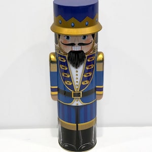 May include: A blue and gold nutcracker tin with a black mustache and beard. The tin is shaped like a standing nutcracker with a tall blue hat and a gold crown.