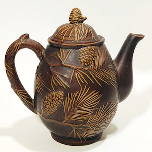 May include: A brown ceramic teapot with a pine cone design. The teapot has a lid with a pine cone finial and a brown handle.