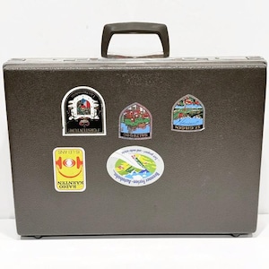 May include: A dark brown vintage briefcase with a black handle and several travel stickers. The stickers feature various designs and text, including "FORSTENIUM", "SALZBURG", and "RADIO KÄRNTEN".