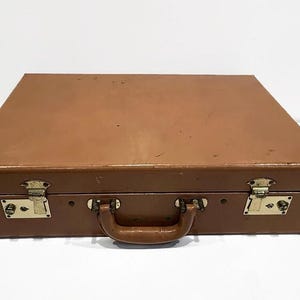 May include: A vintage brown briefcase with a handle and two brass latches. The briefcase is rectangular and has a slightly worn appearance, suggesting age and use. The hardware is gold-colored.