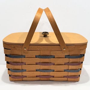 May include: A rectangular woven basket with a wooden lid and handle. The basket is made of light brown woven strips with accents of blue and maroon. The lid has a small brass knob. The handle is made of light brown wood.