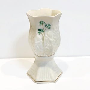 May include: A white porcelain vase with a gold rim and a green shamrock design. The vase has a scalloped top and a pedestal base.