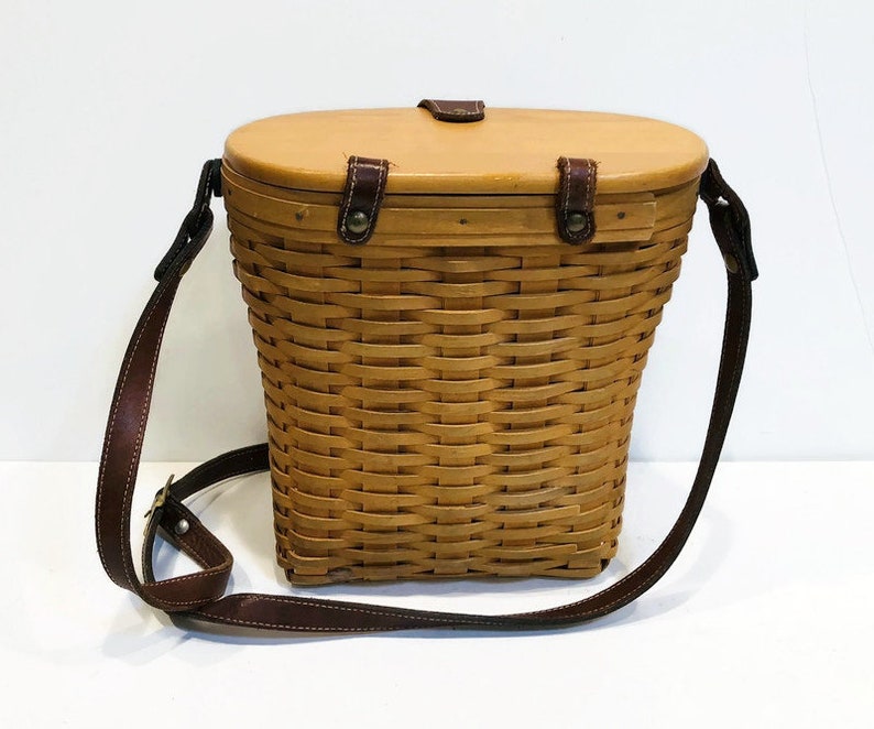 May include: A brown wicker basket with a leather strap and a wooden lid. The basket is oval shaped and has a woven pattern.