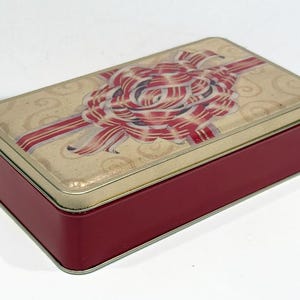May include: A red rectangular tin box with a gold lid. The lid features a colorful illustration of a large bow made of red and white ribbon on a cream background.