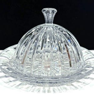 May include: A clear, glass butter dish with a domed lid and a round base. The lid features vertical ribbed detailing and a small, textured handle. The base has a matching ribbed design. The dish is designed to hold butter and is suitable for the kitchen.